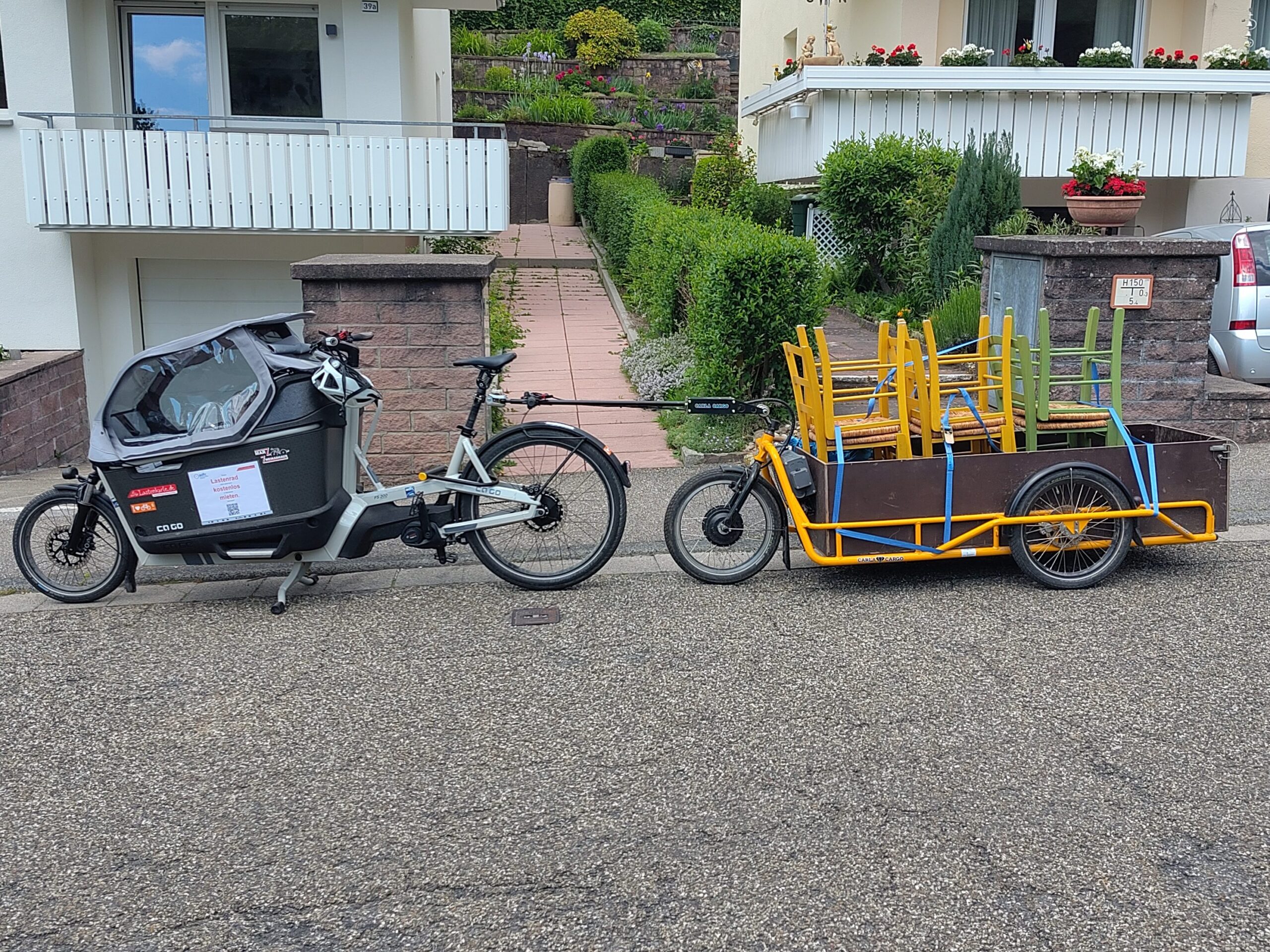Vor zwei Häusern steht das Gespann auf der Straße. Auf dem Anhänger befinden sich sechs mit Spanngurt befestigte Stühle.